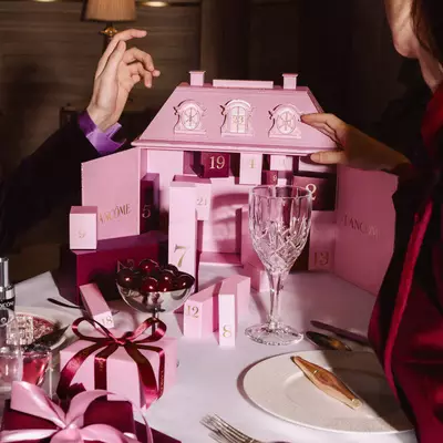 Kutlama sofrasında sofra takımları ve kirazlar eşliğinde pembe bir Lancome Advent Calendar'ı acan eller.